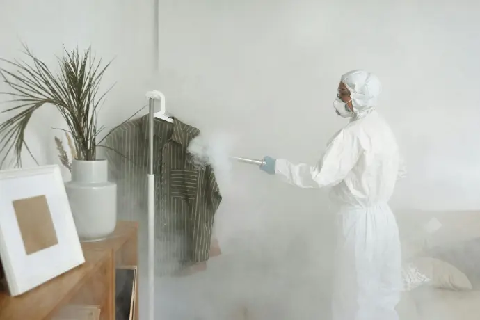 Licensed pest control technician in protective white jumpsuit applying treatment to infested clothing and room surfaces.